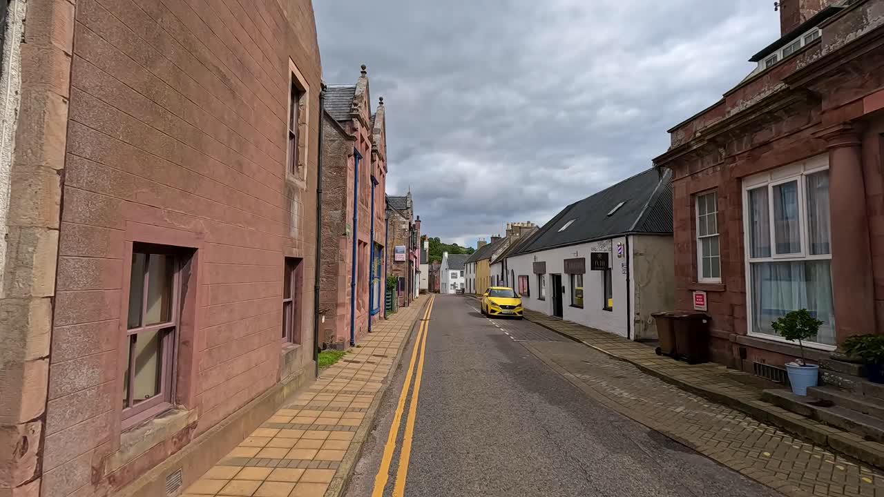 Car drives along narrow street with historic stone houses in a quiet Scottish village, daytime