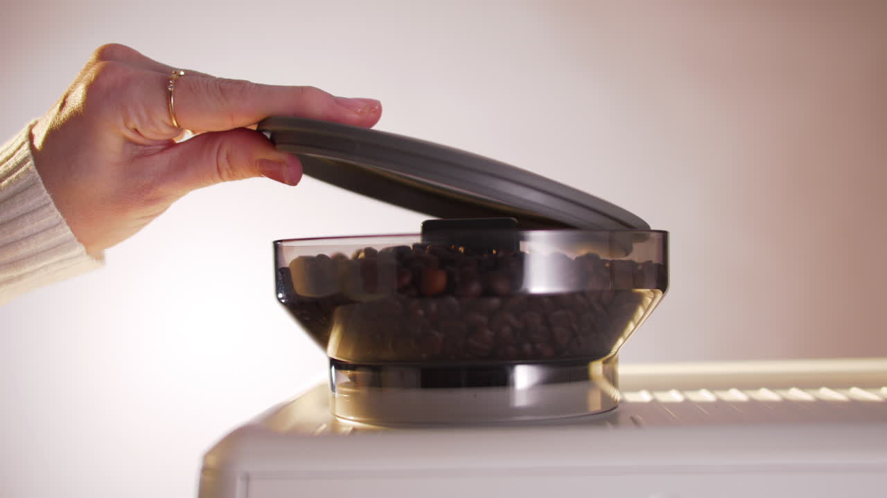 A woman closes the lid of a coffee bean hopper on top of a sleek portafilter espresso machine, emphasizing preparation steps in barista-style coffee making at home or in cafés