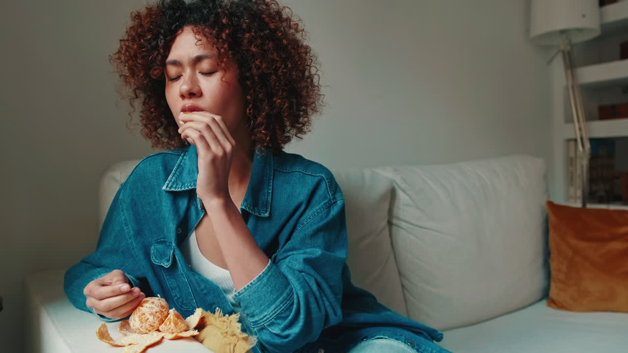 Thoughtful Young Woman Eating an Orange on the Sofa