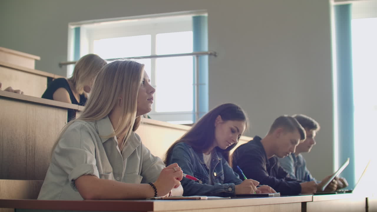 A group of male and female students in a large auditorium of the University listen to a lecture by a Professor.