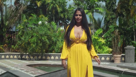 una chica con un suave vestido amarillo fluido caminando por el jardín al aire libre y la piscina de un impresionante castillo.