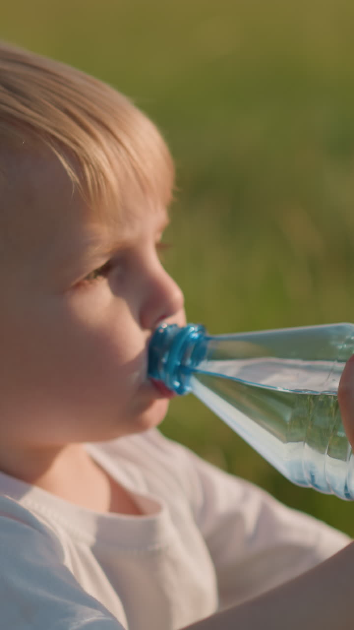 un niño pequeño con una camiseta blanca está sentado en un campo cubierto de hierba, bebiendo agua de una botella de plástico transparente. el niño parece contento y concentrado, disfrutando de un momento refrescante al aire libre iluminado por el sol