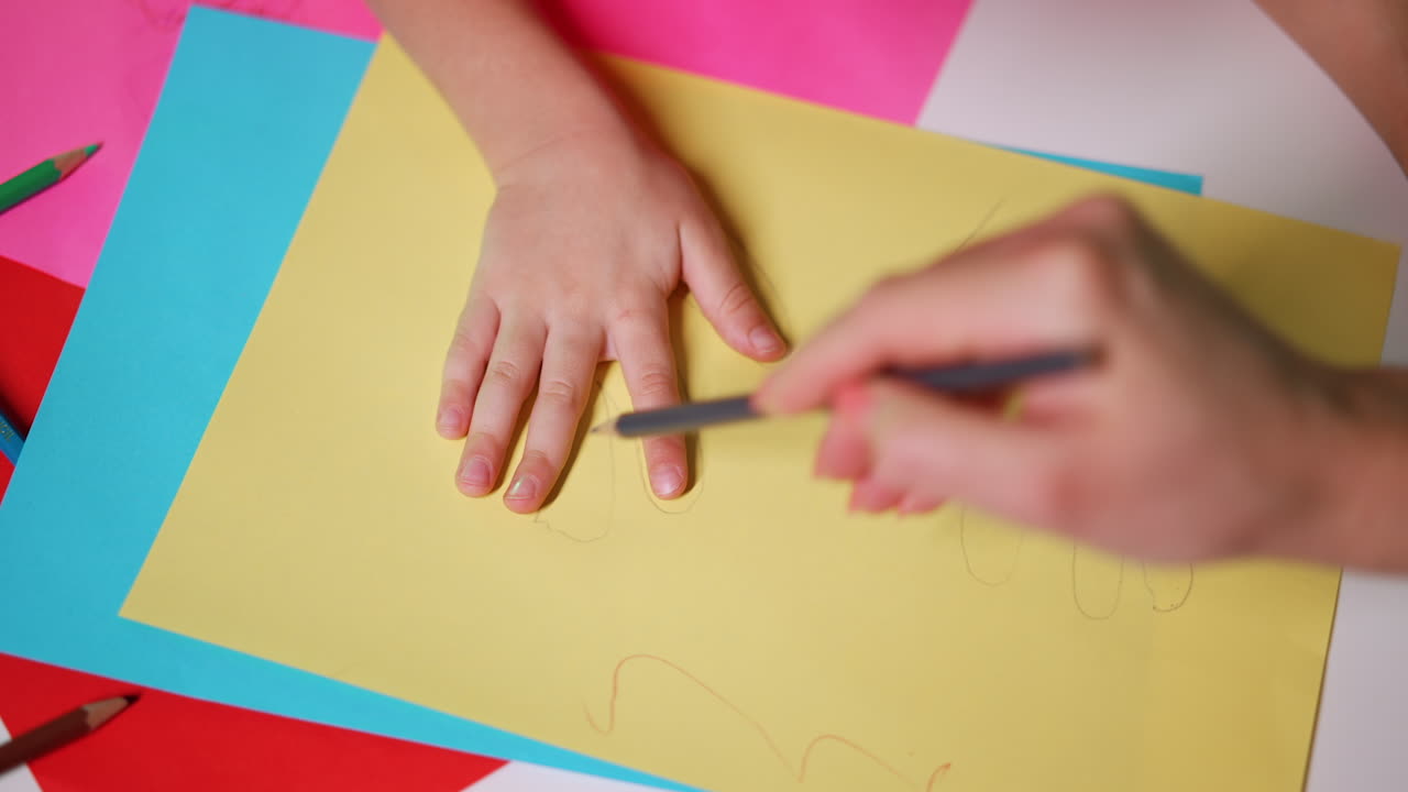 Hands of a little Caucasian baby on the sheet of paper. Mom's hand drawing lines outlining child's fingers. Close up.