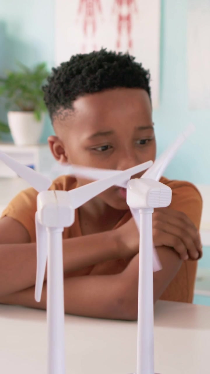 Vertical video: Reaching youth in orange tee spinning and inspecting two turbines on table in study