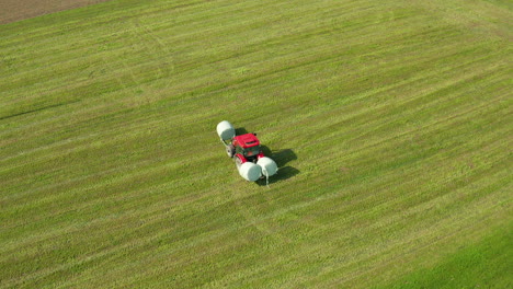 Red-tractor-picking-up-bales-of-grass-on-green-meadow,-farming-and-agriculture-concept,-aerial-view