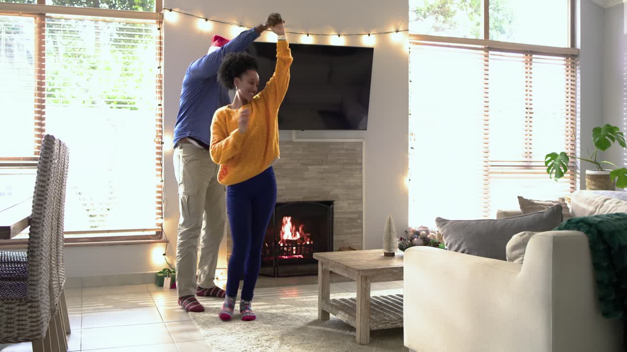 Diverse couple receiving holiday gift, dancing twirling by lit fireplace amid lights with Santa hat