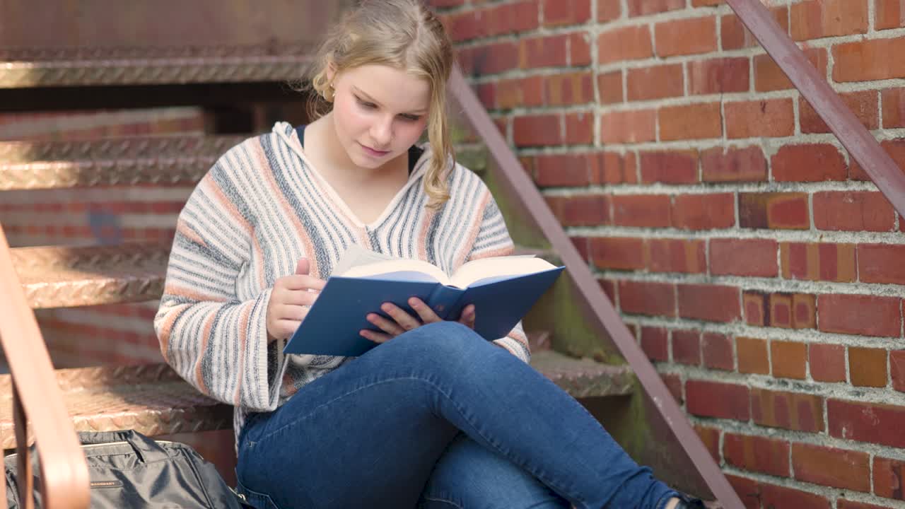 Studentin liest draußen auf den Stufen ein Buch und blättert eine Seite ihres Lehrbuchs um.