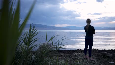 young woman standing alone at calm waterfront lake with mountain range in background on cloudy day, handheld pan slow motion