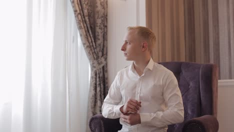 Handsome-groom-man-dresses-in-wedding-morning-and-fixes-his-buttons-on-a-white-shirt-sleeve,-indoors