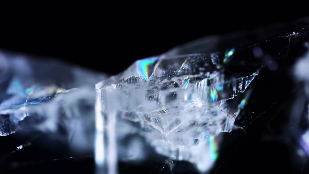 Extreme close-up of fractured translucent crystalline shard against deep black background, with changing cool light, sharp ridge detail, layered internal fissures, iridescent glow, and fine filament