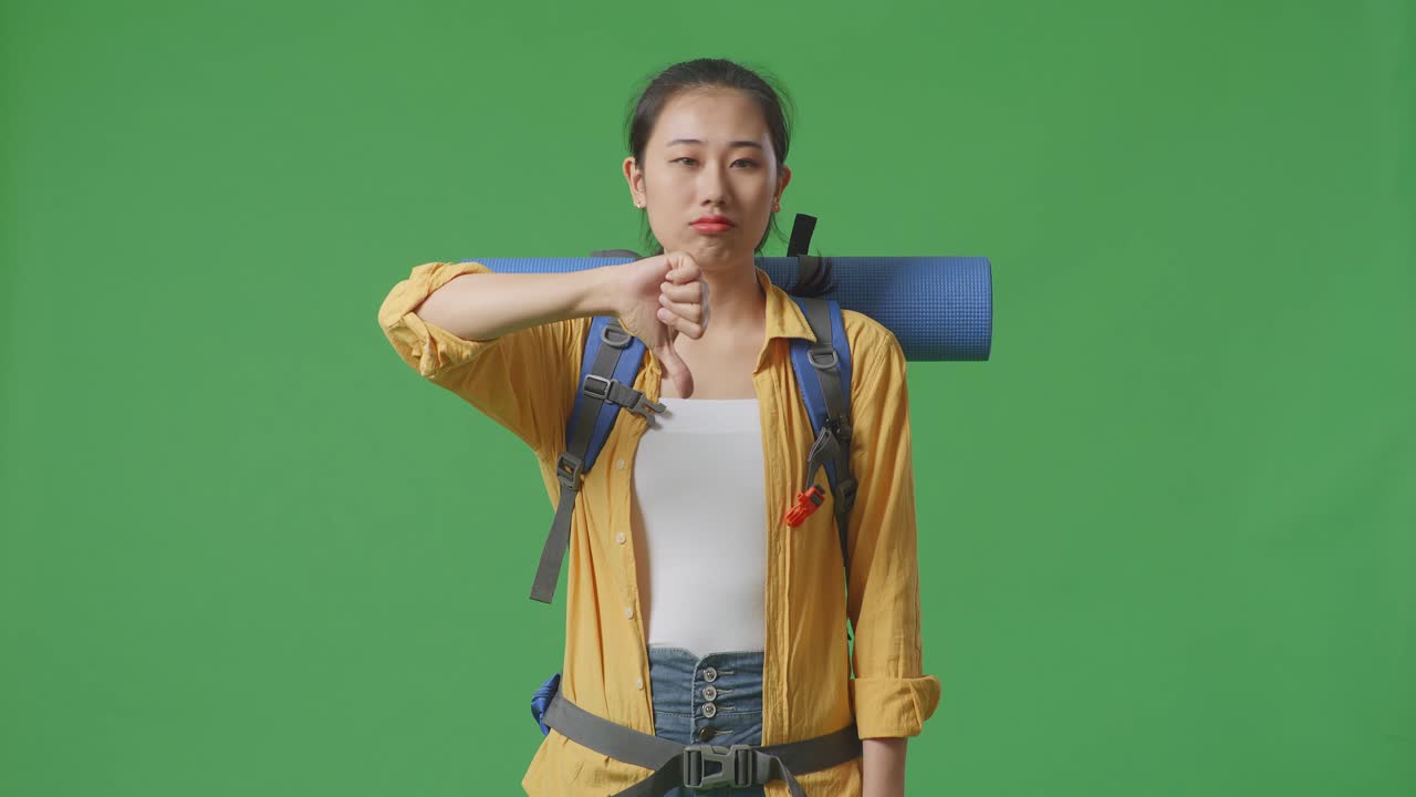 아시아 여성 하이커가 산악 배을 들고 스튜디오에서 녹색 스크린 배경에 서서 엄지손가락을 내려놓은 몸짓을 보여줍니다.