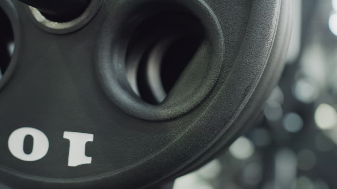 Close up view of textured black weight plate wheel mounted on gym machine, showcasing circular grip hole, rubber surface grain, blurred equipment in background, emphasizing fitness gear detail