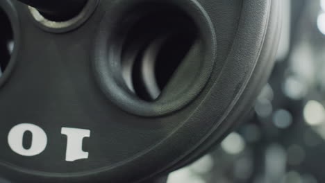 close up view of textured black weight plate wheel mounted on gym machine, showcasing circular grip hole, rubber surface grain, blurred equipment in background, emphasizing fitness gear detail