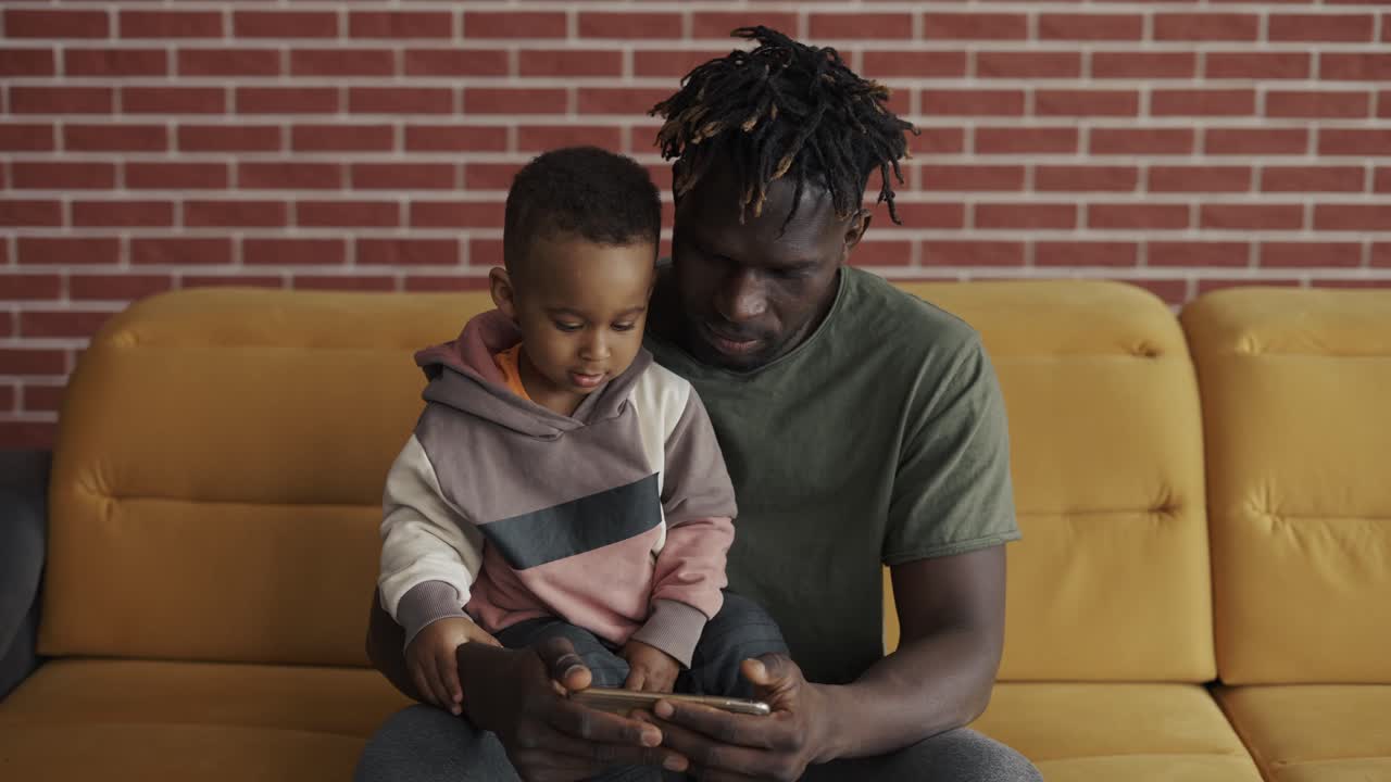 Portrait of african american father sitting with son on sofa watching cartoon on smartphone