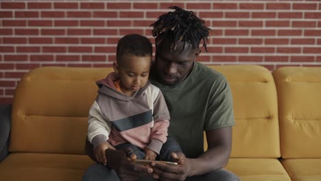 portrait of african american father sitting with son on sofa watching cartoon on smartphone