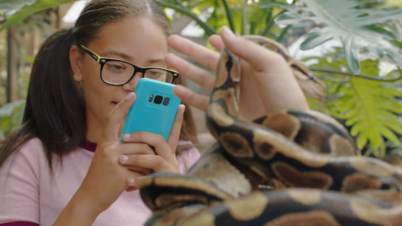 chica joven sosteniendo una serpiente con un amigo tomando fotos usando un teléfono inteligente compartiendo una excursión al zoológico en las redes sociales aprendiendo sobre reptiles en el santuario de vida silvestre 4k
