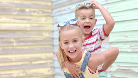 Portrait-of-happy-caucasian-sister-carrying-her-brother-with-sunglasses-over-beach-house