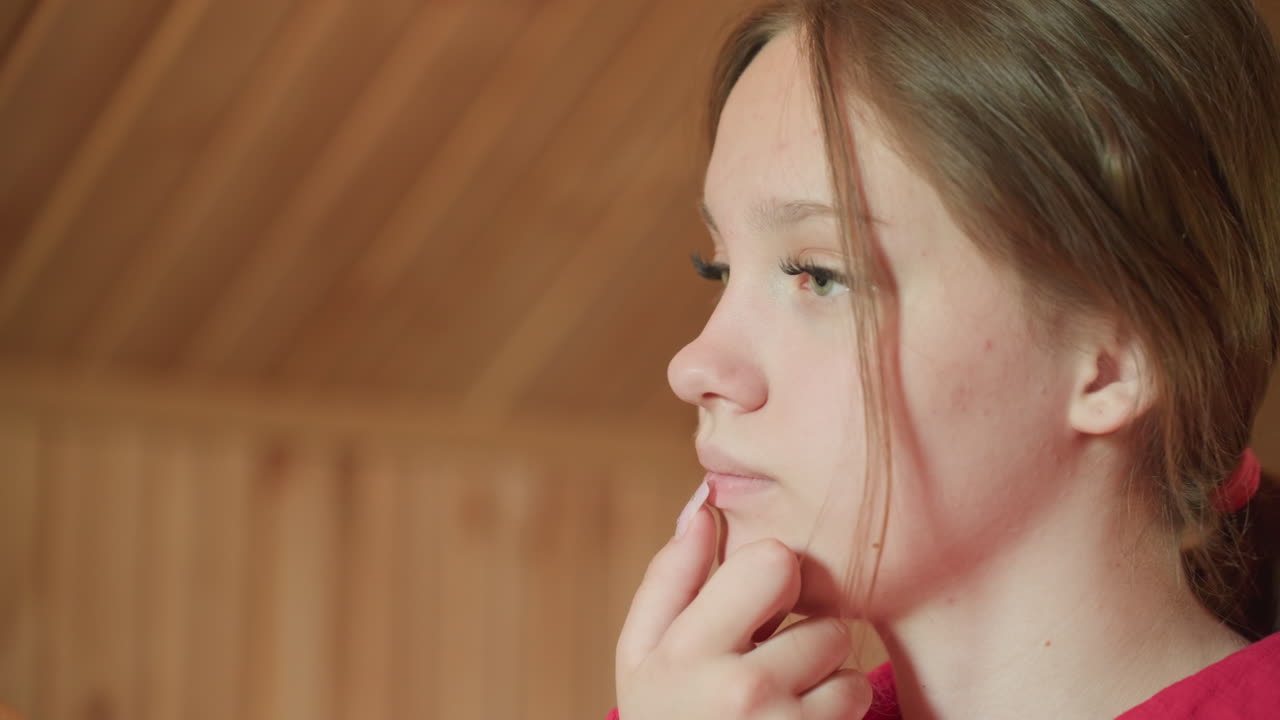 side profile teenage girl in red shirt touching chin with thoughtful expression in wood paneled attic room while watching friend operate smartphone, quiet intimate portrait capturing pensive mood