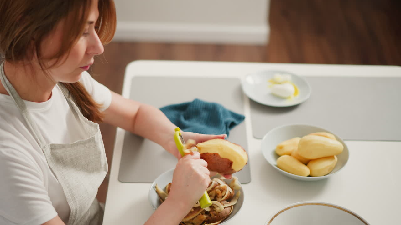 Chef peels potato with head tilted left while seated at kitchen table, surrounded by peeled potatoes, potato skins, and a kitchen cloth, in bright modern kitchen with visible egg plate nearby