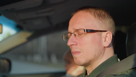 una toma de primer plano de un hombre con gafas y una chaqueta verde, sentado en el asiento del conductor de un coche. la expresión en su rostro refleja el enfoque y la contemplación, mientras mira hacia adelante