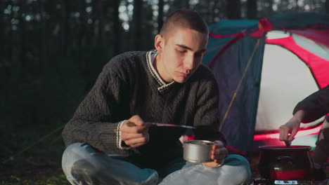 couple seated outdoors near camp tent man eating can food while wife stirs pot on portable stove camping forest nature picnic leisure casual clothing smiling happy lifestyle