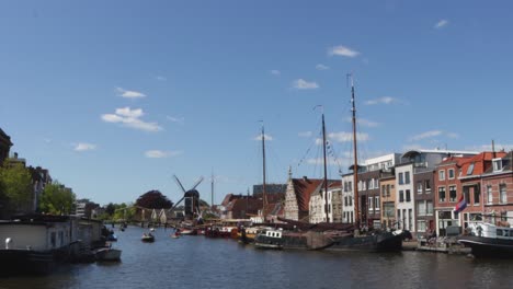 maritime traffic, docked ships and houseboats on a historical canal in the netherlands, with old windmill and bridge in the background, on a sunny day