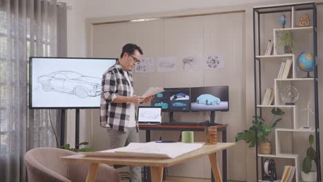 asian male looking at car design sketch paper in his hands and walking around while working in the studio with tv and computers display 3d electric car model