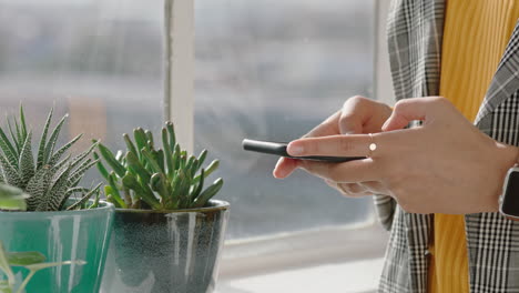 Cerrar-Las-Manos-De-Una-Mujer-Usando-Un-Teléfono-Inteligente-En-Casa-Enviando-Mensajes-De-Texto-Escribiendo-Mensajes-De-Correo-Electrónico-Navegando-En-Las-Redes-Sociales-En-Línea-De-Pie-Junto-A-La-Ventana