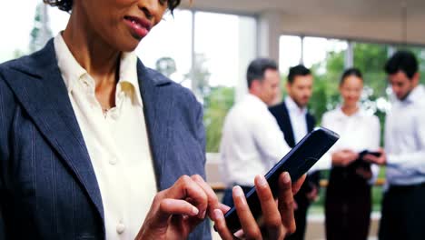 ejecutiva de negocios enviando mensajes de texto en el teléfono móvil mientras sus colegas interactúan en segundo plano
