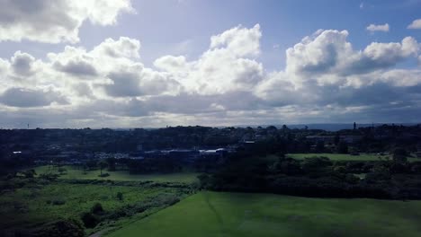 Drone-Ascendiendo-Sobre-Un-Campo-Deportivo-Verde-Con-Vistas-A-Un-Centro-Comercial-Con-Nubes-En-El-Cielo