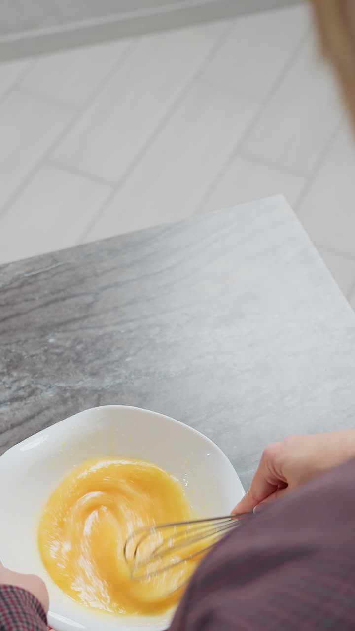 Rear view of woman with golden hair wearing plaid gown as she stirs egg in a bowl, soft lighting highlights her posture and focus, with a blurred countertop and kitchen setting