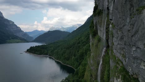 Beautiful-waterfall-cascading-from-rocky-Alps-with-Klontalersee-lake-backdrop