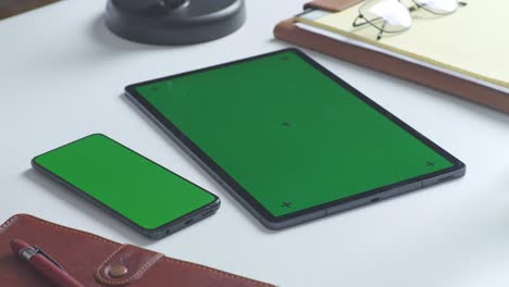 top down view of a laptop computer with mock up green screen chromakey display on a wooden office desk next to notebook with pens, glasses, and a glass of water. slow zoom out, close up