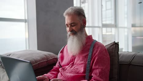 a serious and happy elderly man with gray hair and a lush beard in a pink shirt sits on a sofa in a modern apartment and works on a laptop