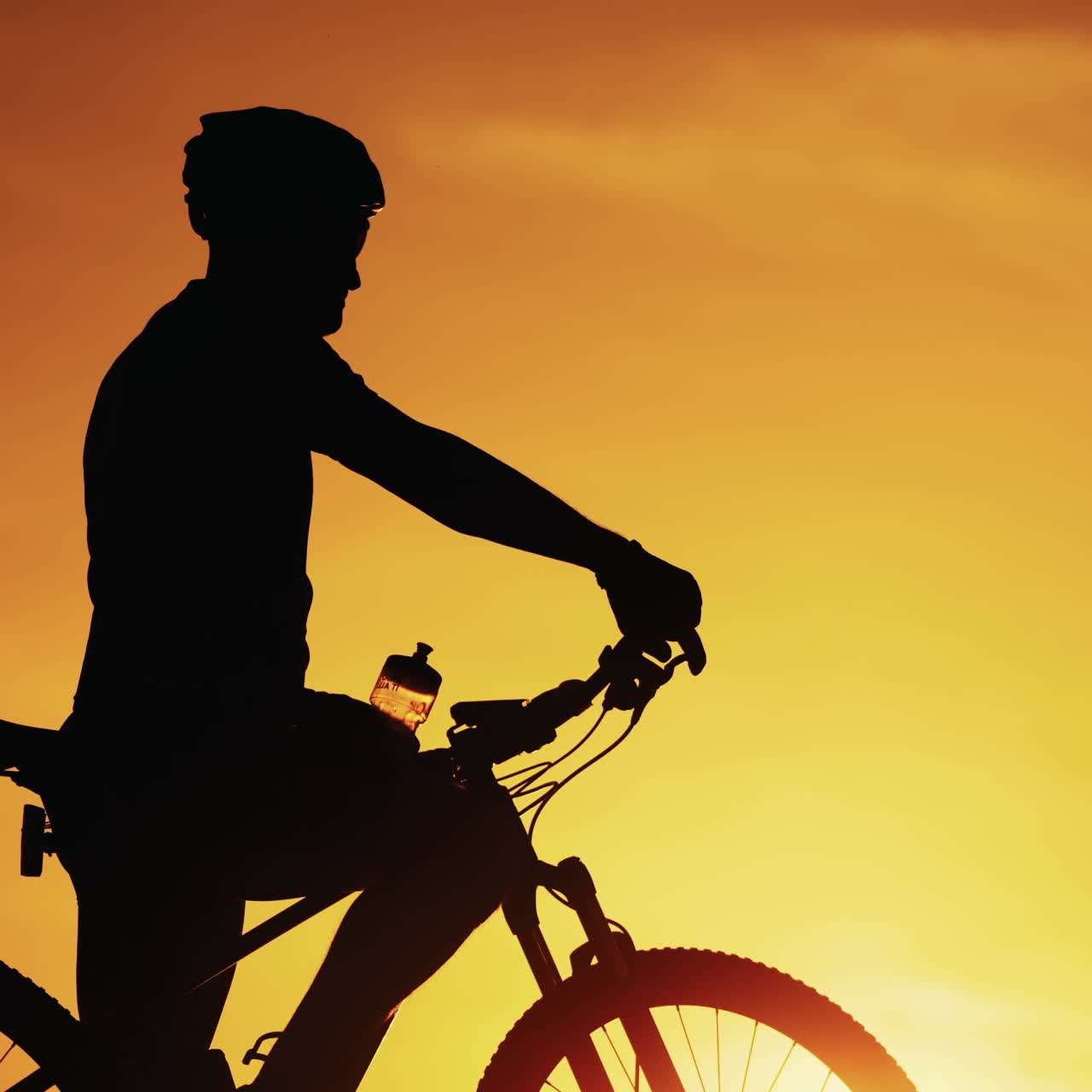 Silhouette of a cyclist man is drinking water after intensive cycling training at sunset. Biker drinks water from bottle and puts it into a bike on the natural golden sun setting background.