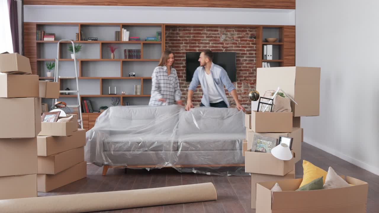 새로운 아파트의 소파에서 보호 개를 제거하는 젊은 부부 - 이사하는 날 행복한 주택 소유자 - 부동산 및 임대 개념 - 젊은 가족을위한 대출 모기지