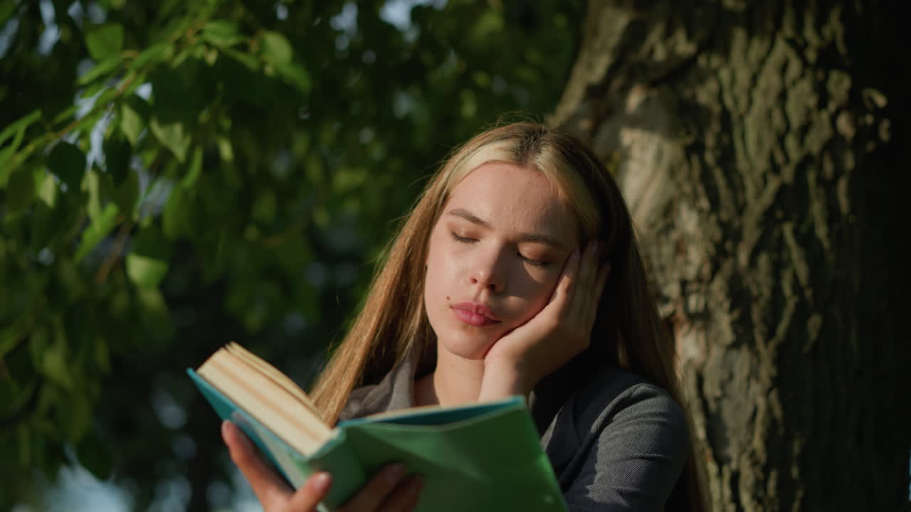 dama sentada bajo un árbol con la cabeza apoyada en su mano izquierda, leyendo un libro, las páginas volviendo en el viento, mira hacia la distancia con aspecto de mal humor, la luz del sol ilumina su rostro, el fondo suavemente borroso