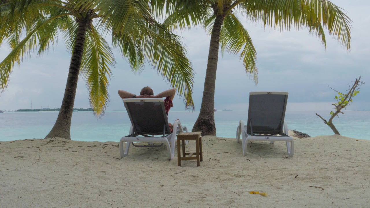 Un vacanziere si riposa con le mani dietro la testa su un lettino prendisole accanto a un secondo lettino vuoto, di fronte a una calma laguna turchese da una spiaggia delle Maldive ombreggiata da palme, in un'inquadratura ampia e statica