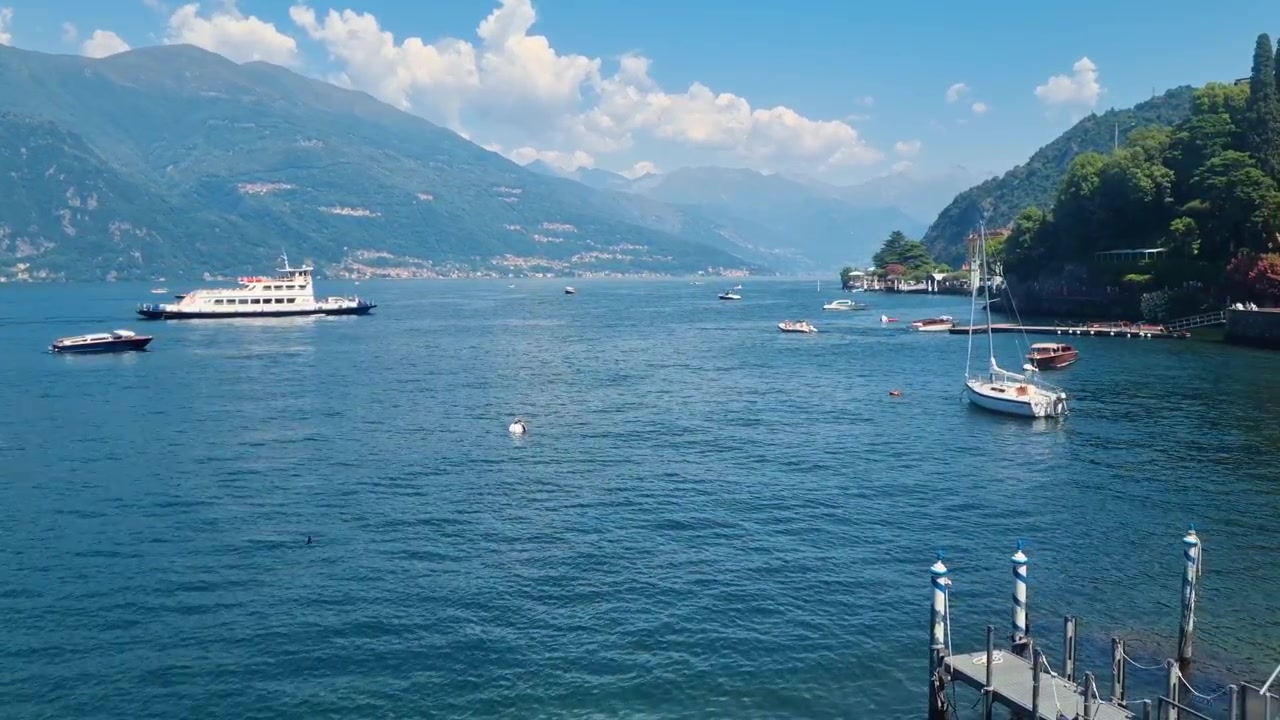 Panorama estivo pittoresco del Lago di Como a Varenna, Italia, dove piccole barche riposano e un traghetto si muove su acque cristalline sotto cieli blu. Una destinazione lacustre senza tempo.