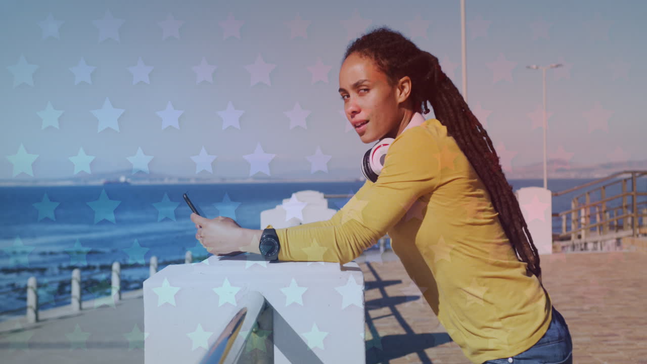 múltiples iconos de estrellas contra una mujer afroamericana que usa un teléfono inteligente en el paseo marítimo