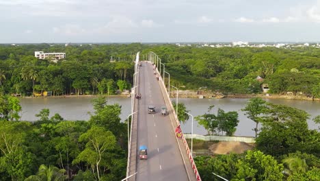 aerial descending view of viaduct bridge over river with forested riverbank, vehicles driving by, bangladesh