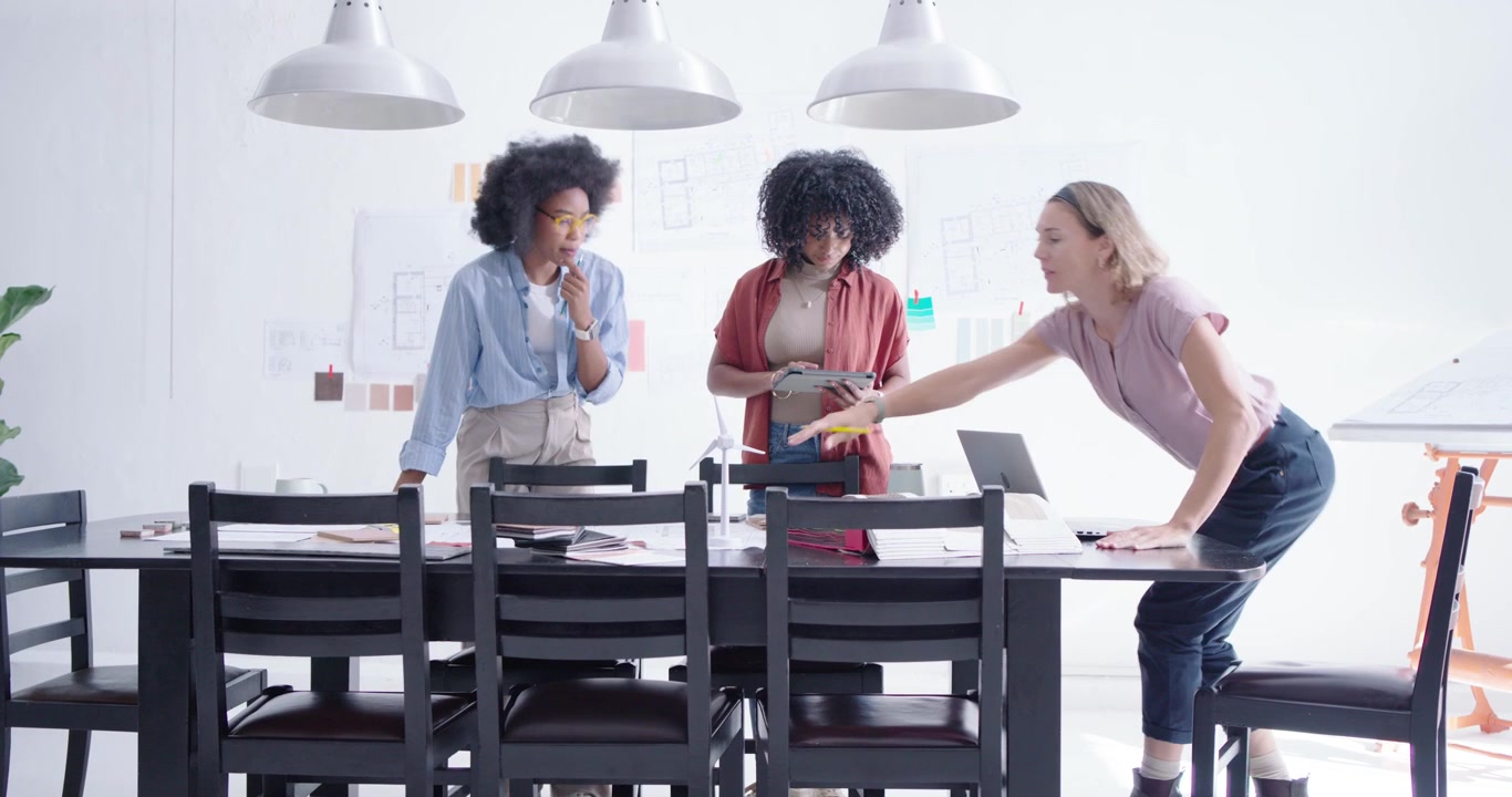 Diverse female team reviewing layouts at table, moving swatches to wall, choosing palette on laptop