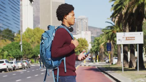 hombre afroamericano en la ciudad, usando un teléfono inteligente, con auriculares y mochila cruzando la calle