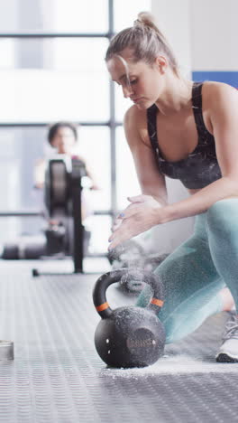 vertical video of confident caucasian woman chalking hands before lifting kettlebell weight at gym
