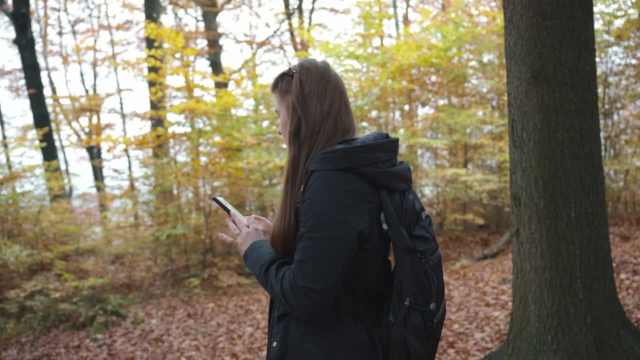 Girl with long hair is using the phone in the middle of the forest. Autumn among trees and fallen leaves. Walking and exploring on a nice day