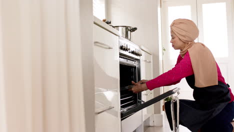 biracial woman in hijab putting tray into oven in kitchen at home with copy space, slow motion