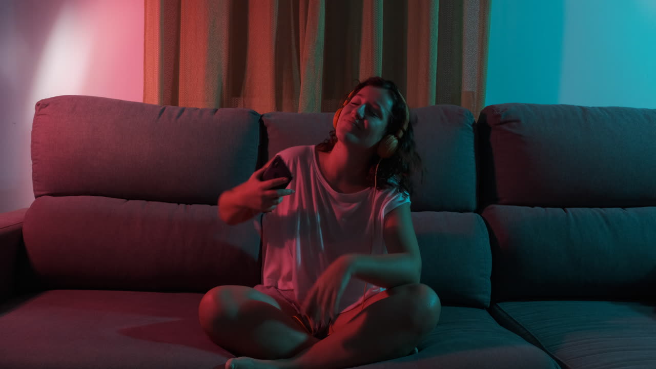 Young woman alone listening to music with headphones at home, colorful blue and red lights