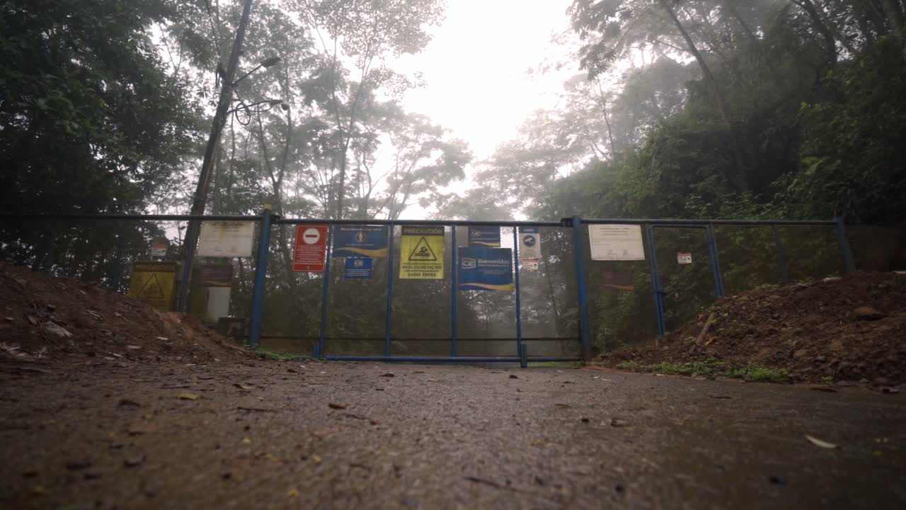 Metal gate with several warning and informational signs, blocking a muddy trail. The scene is set in a foggy, rainy environment, with trees and lush foliage visible in the background