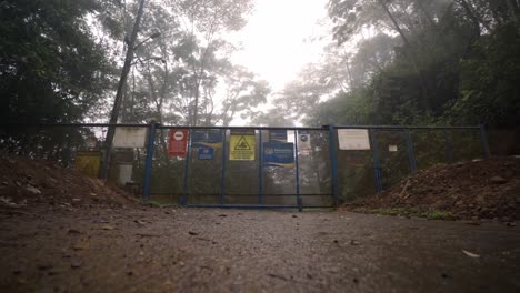 metal gate with several warning and informational signs, blocking a muddy trail. the scene is set in a foggy, rainy environment, with trees and lush foliage visible in the background