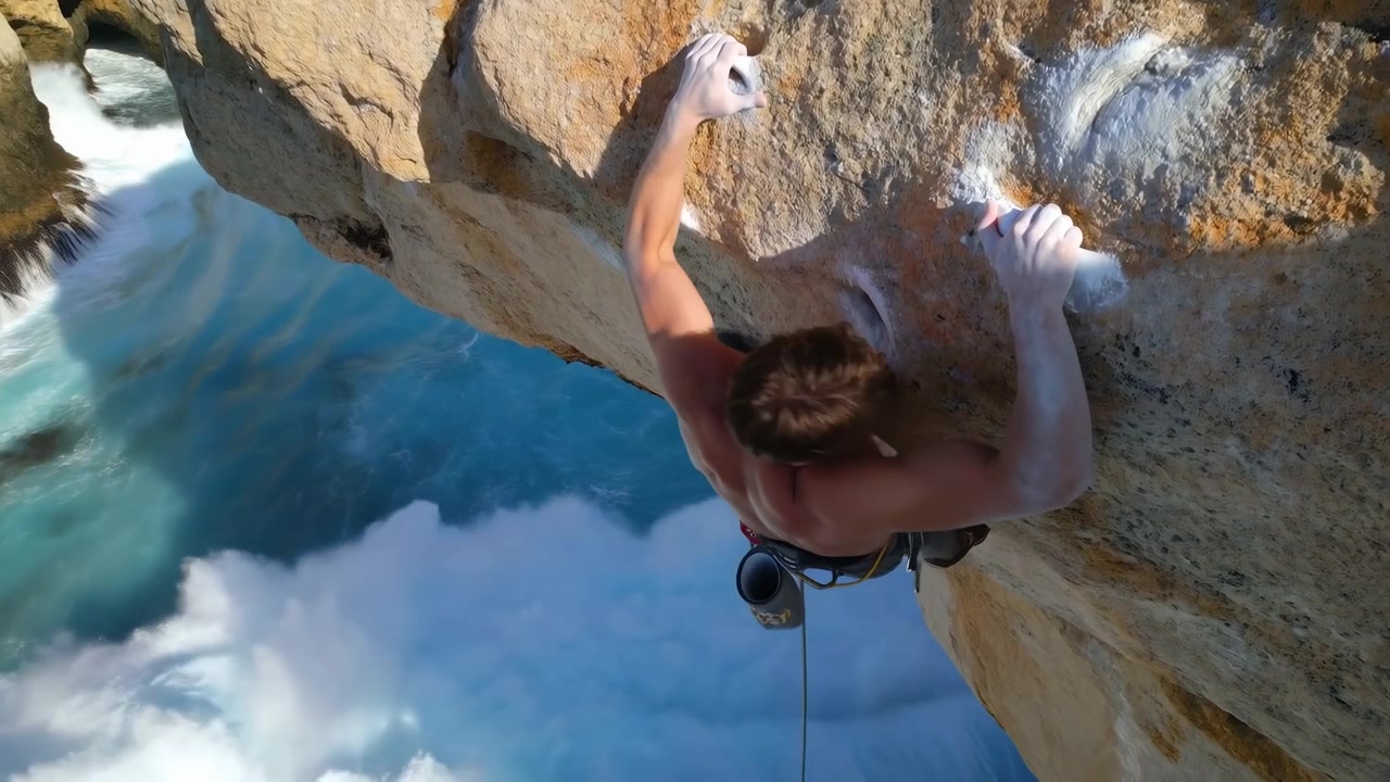A shirtless climber is seen from above on the right side of the frame, stretching across a rugged tan cliff with one arm extended while bright blue water